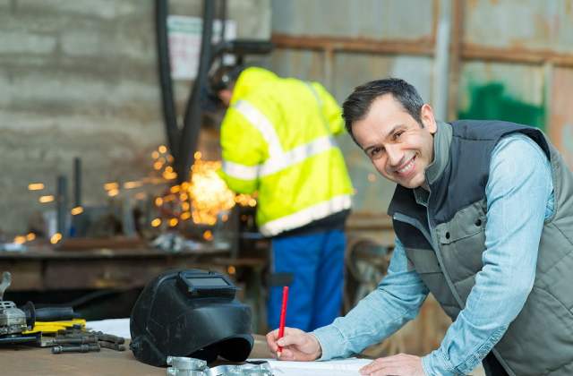 Photograph of man in a workshop, writing, with welder or steel worker in the background